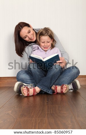 Education at home for young primary school girl with mother teaching her to read a book. Both wearing denim jeans and hoodies sitting cross legged on the floor.