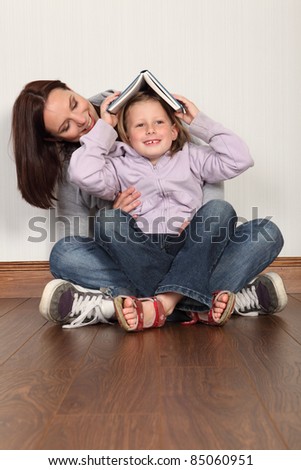Education at home for young primary school girl having fun putting book on her head with mother trying to teach her to read. Both wearing denim jeans and hoodies sitting cross legged on the floor.