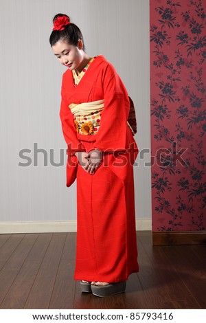 Respectful bow by beautiful young oriental model in red Japanese kimono robe garment complete with obi sash and kanzashi hair flower.