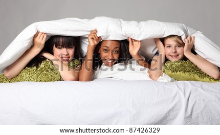 Sleepover party bedtime fun for three happy beautiful teenage girl friends, a mixed race african american, oriental Japanese and blonde caucasian school mates laughing under bed covers.