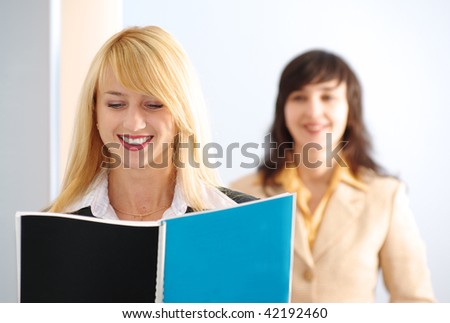 Blonde girl reading the plain covered book while brunette is approaching, in the office