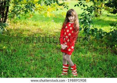 Adorable kid girl in red sweater dress and stripe rainboots having fun outdoors in the sunny autumn day