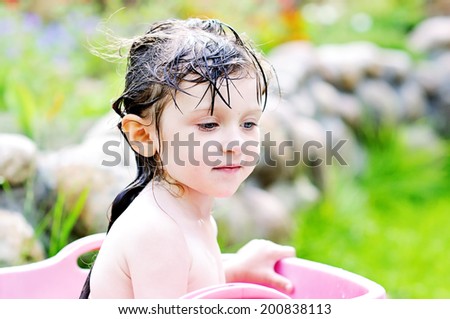 Adorable toddler girl with brunette hair having bath and playing with water in the pink washbowl outdoors  in the garden on the hot summer day