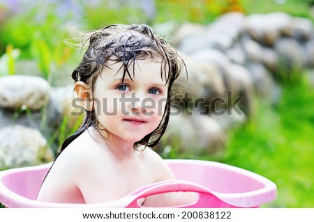 Adorable toddler girl with brunette hair having bath and playing with water in the pink washbowl outdoors  in the garden on the hot summer day