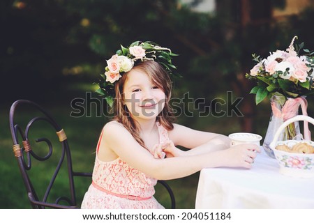 Beauty kid girl in flower wreath having tea in the garden