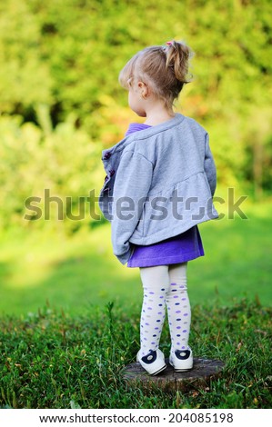 Adorable toddler girl with curly blond hair in knit purple dress and grey jacket playing outdoors on beauty sunny day