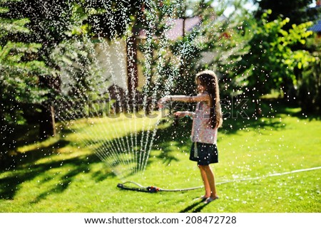 Nice kid girl with long hair has fun with garden sprinkler playing with water splashes in the backyard on a sunny hot summer  evening.