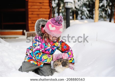 Beauty little girl in colorful winter jacket and hat plays with a furry cat in deep snow on nice winter day