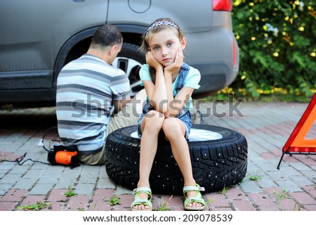 Unhappy kid girl sitting on the wheel and waiting her father to change broken wheel during the family trip