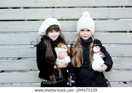 Two adorable kid girl friends in white hats and pattern black jackets has fun outdoor at the winter day