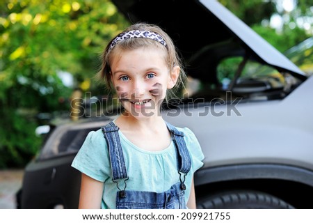 Portrait of cute little girl with dirty face helps her father to change wheel on their family car on warm day in the yard