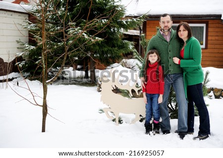 Happy family of three in the red green sweaters outdoor near the house at the snow winter day