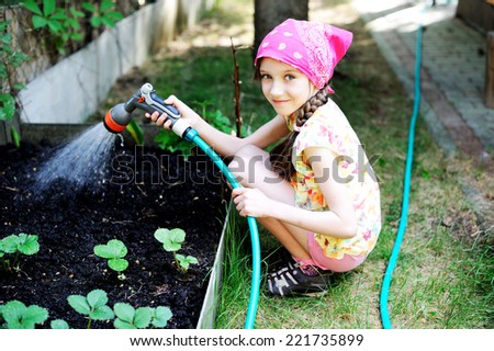 Aadorable brunette kid girl, watering the plants, from hose spray in the garden at the backyard of the house on a sunny summer evening