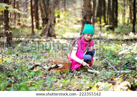 Adorable kid girl in pink vest, white sweater, green hat and boots with basket picking mushrooms in autumn forest at sunny day