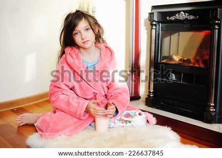 Adorable kid girl in pajama sitting near fireplace with glass of milk