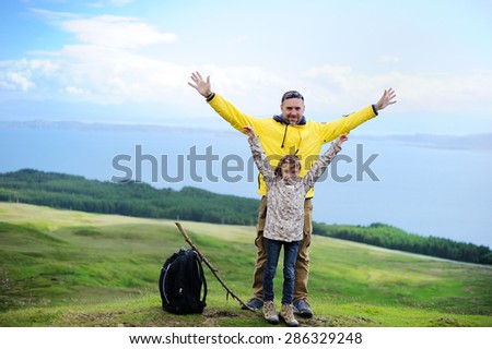Hiking family adventure on mountain trek: beauty kid girl and her  active father in jackets and special boots having rest on green lawn with mountain lake view: Scotland, Skye