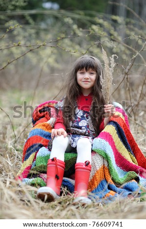 Adorable brunette little girl with very long hair in colorful dress and  red rain boots in the field with colorful stripe blanket