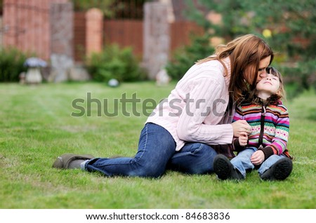 Young brunette mother kisses her child daughter sitting on the green lawn