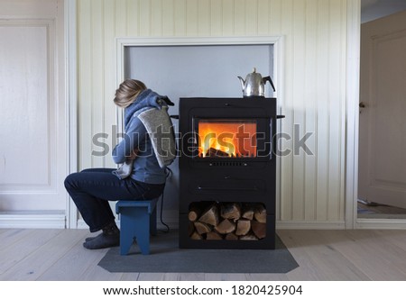 Woman warming herself by the side of metal stove; fire burning in stove, firewood under stove and coffee pot on stove. 