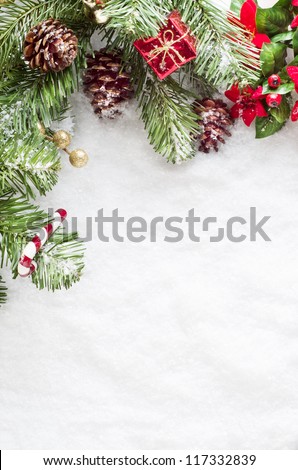 A Christmas border to left and top of frame consisting of artificial foliage, real pine cones and decorative ornaments, sprinkled with snow on a fake snow background.   Snow provides copy space.