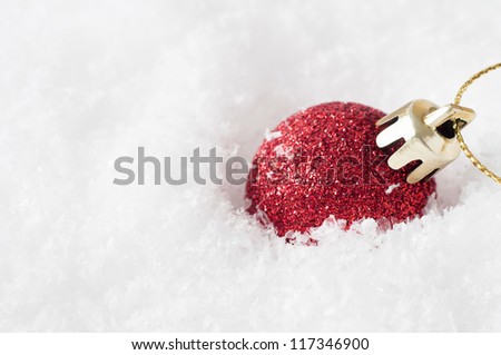 A solitary shiny red Christmas bauble, coated in glitter with gold clasp and string, sunk into artificial white snow and leaning to the right.  Copy space to left.