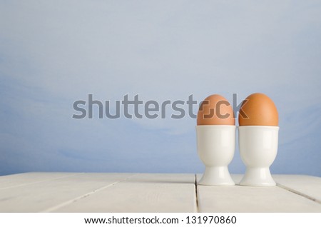 Two fresh brown eggs in egg cups on an old, cream painted wood plank farmhouse table, with painted blue sky effect background.