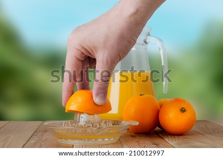 A hand squeezing juice from an orange on a manual glass squeezer.  Set on a wooden planked table with a group of three oranges and a glass jug of juice.  Outdoor background of soft foliage.