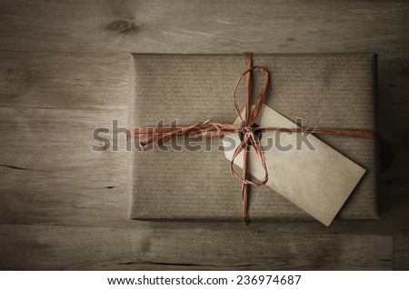 Overhead shot of a gift box with a grungy parcel tag for a label, wrapped in brown paper with a raffia ribbon tied to a bow on wooden planked table.  Vintage appearance with dark tones and  vignette.