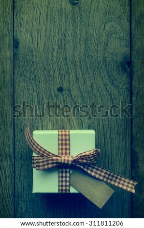 Overhead white box on old wood plank table, tied to a bow in red and cream gingham ribbon, with vintage effect parchment style label left blank for copy space.  Cross processed for retro appearance.