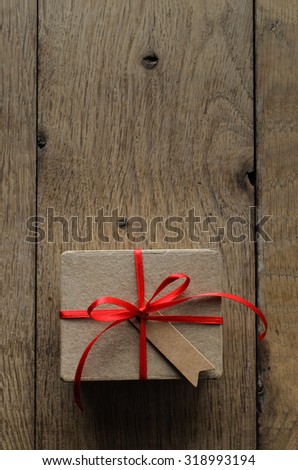 Overhead shot of a simple brown gift box on an old oak wood planked table, tied to a bow with red satin ribbon, with a blank vintage style parcel tag facing upwards.