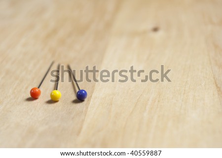 Close up of three steel pins with colorful plastic heads (orange, yellow and purple) lying on a birch wooden table in lower left frame, with empty table space to the right and above.