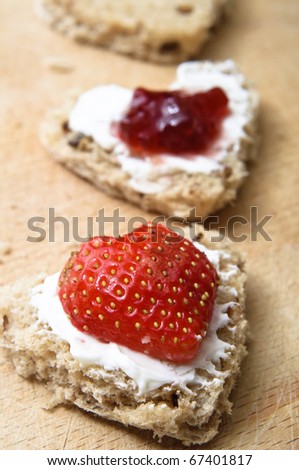 A vertical row of heart shaped granary bread snacks on a wooden chopping board.  Toppings of cream cheese, cut strawberry and jam.