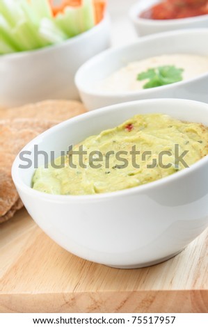 Guacamole, hummus and salsa dips in white bowls on a wooden board with wholemeal pitta bread and crudites cropped at left frame.