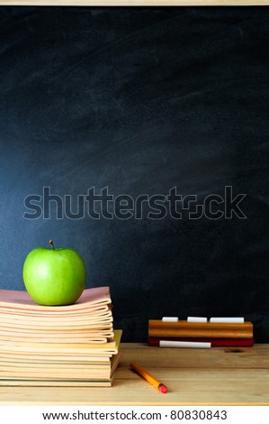 A school chalkboard and teacher\'s desk with stack of exercise books and an apple. Copy space on blackboard.  Portrait (vertical) orientation.