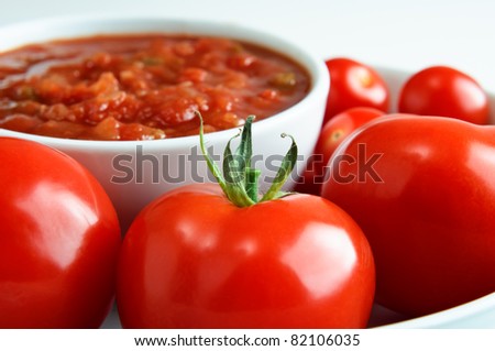 Close up (macro) of bright red tomatoes, surrounding a bowl of salsa in soft focus in the background.  Horizontal (landscape) orientation.