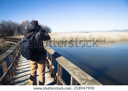 Man watching nature with binoculars, in a wood bridge over the water.