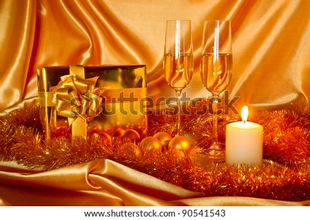New Year (Christmas) still life in golden tones with gift box, candle, satin, Christmas-tree decorations and two galsses of champagne