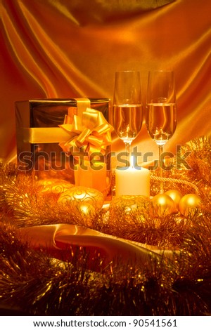 New Year (Christmas) still life in golden tones with gift box, candle, satin, Christmas-tree decorations and two galsses of champagne