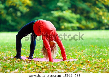 Young happy woman doing yoga in autumn park outdoors. Sport fitness and exercising in nature. Bridge yoga pose over green forest fall background