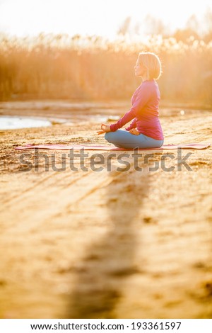 Young woman doing yoga near lake outdoors, meditation. Sport fitness and exercising in nature, autumn sunset