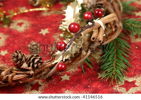 Christmas wreath with golden holly leaves, cones, red berries, cinnamon and twig spruce on red textile with gold stars. Shallow DOF