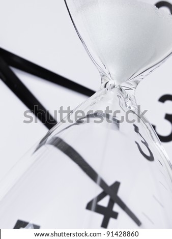 Time running out: White sand falling inside hourglass, with analog clock in background (focus on neck of hourglass), shallow depth of field