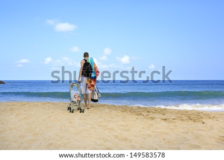 Hands full during vacation with baby. Young father is dragging stroller with little baby on the sandy beach near ocean.