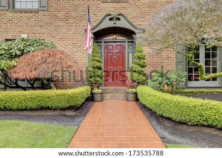 Brick house with large windows and exterior shutters, red wood door, stoned walkway, trim hedge