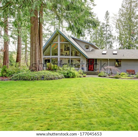 Tile roof countryside house with glass wall, porch and garage. Green lawn together with fir trees and flower bed make curb appeal stand out.