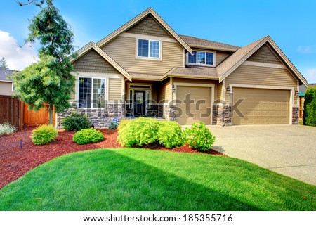 Clapboard siding house  with stone trim. View of  entrance porch, beautiful flower bed with green lawn, bushes and fir tree