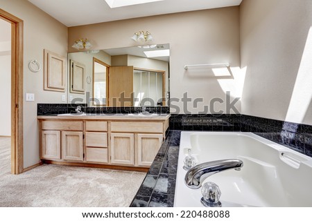 Bright bathroom interior with carpet floor, white bath tub with black granite tile trim. Vanity cabinet with granite top and large mirror