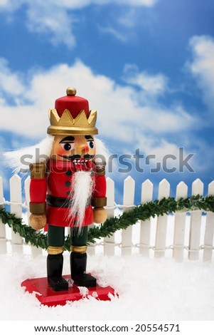 Nutcracker sitting on snow with white fence and green garland on a sky background, nutcracker
