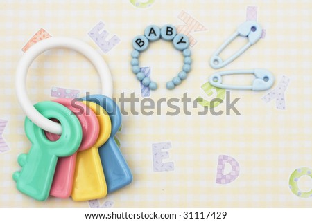 Key shaped baby rattle with pink bracelet and diaper pins sitting on a yellow alphabet background, baby rattle