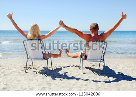 Positive young couple sitting on the beach chair on the sea beach up their hands and looking at see surface